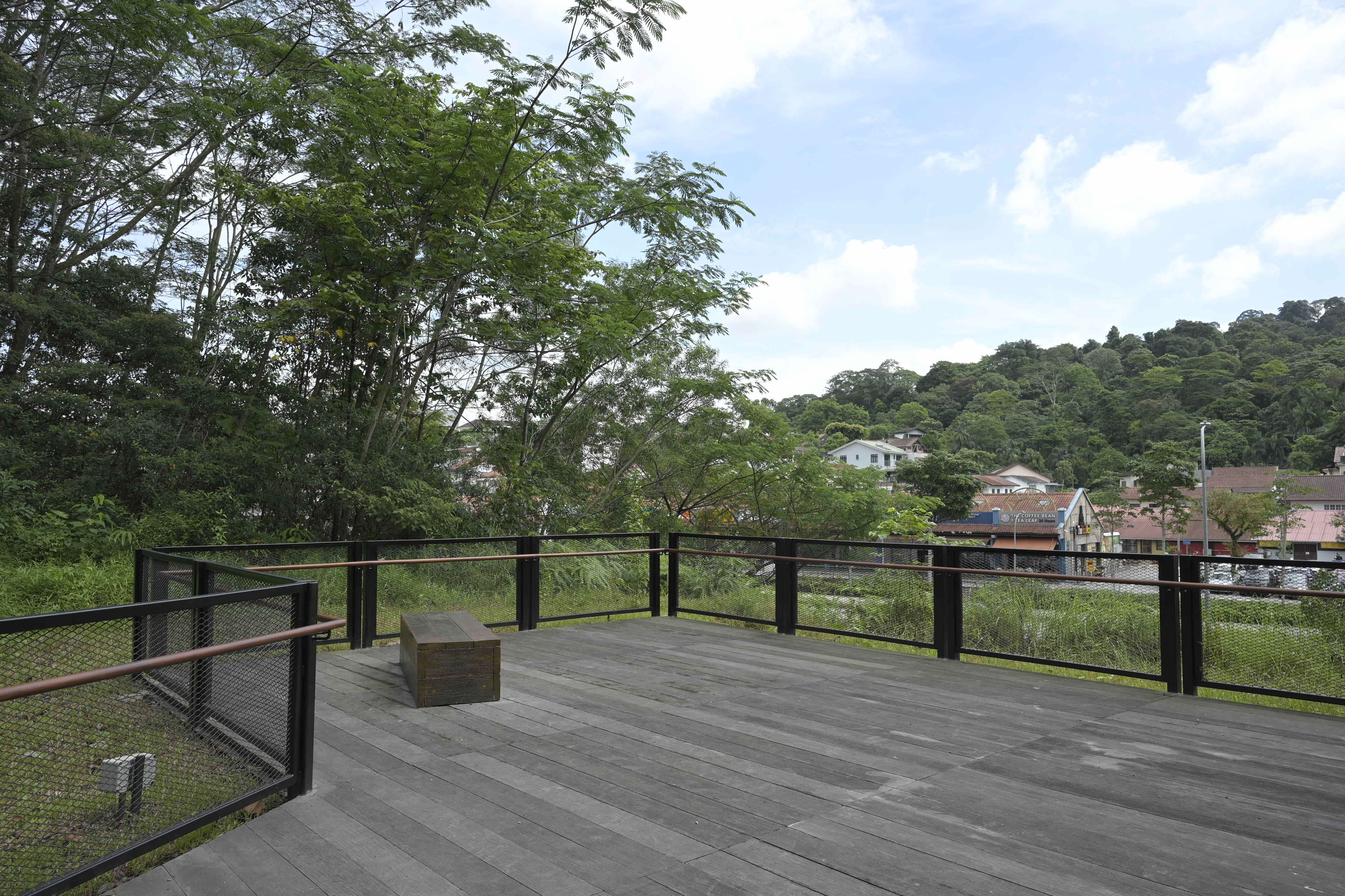A wooden deck with a bench is surrounded by trees, overlooking buildings in a hilly area under a partly cloudy sky.